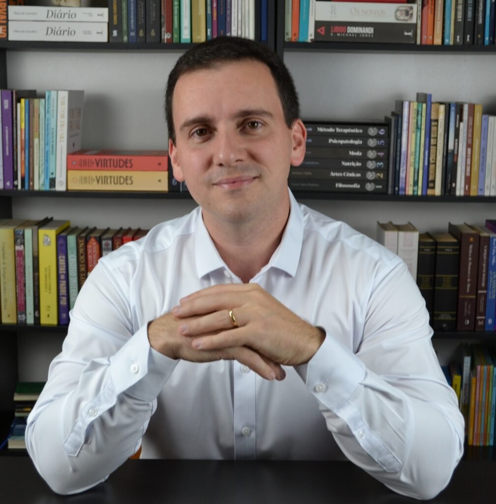 Bruno Vezzoli, autor do Axis Framework, de camisa branca e mãos unidas à mesa, diante de uma estante de livros